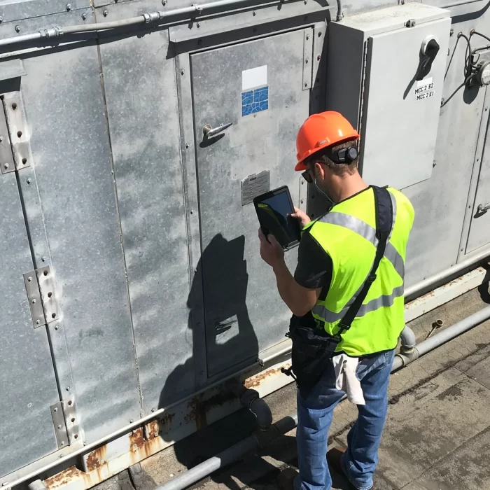 A person wearing a high-visibility vest and orange hard hat operates a tablet while standing next to industrial equipment on a rooftop.