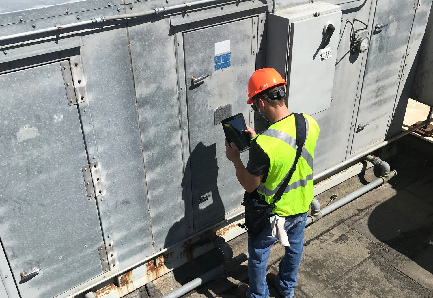 A person wearing a high-visibility vest and orange hard hat operates a tablet while standing next to industrial equipment on a rooftop.