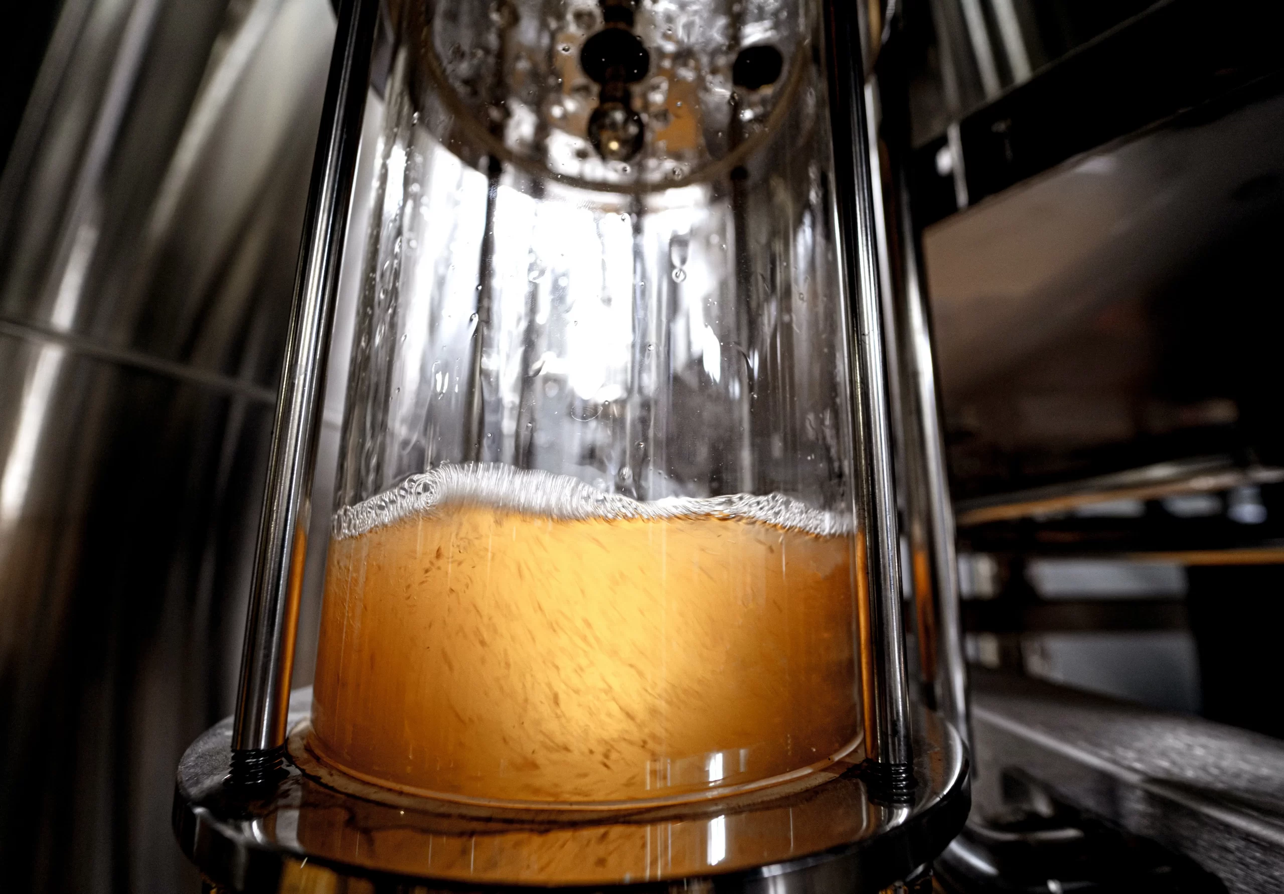 Close-up view of a glass chamber holding amber-colored liquid with visible bubbles. The chamber is part of a shiny metal industrial apparatus.