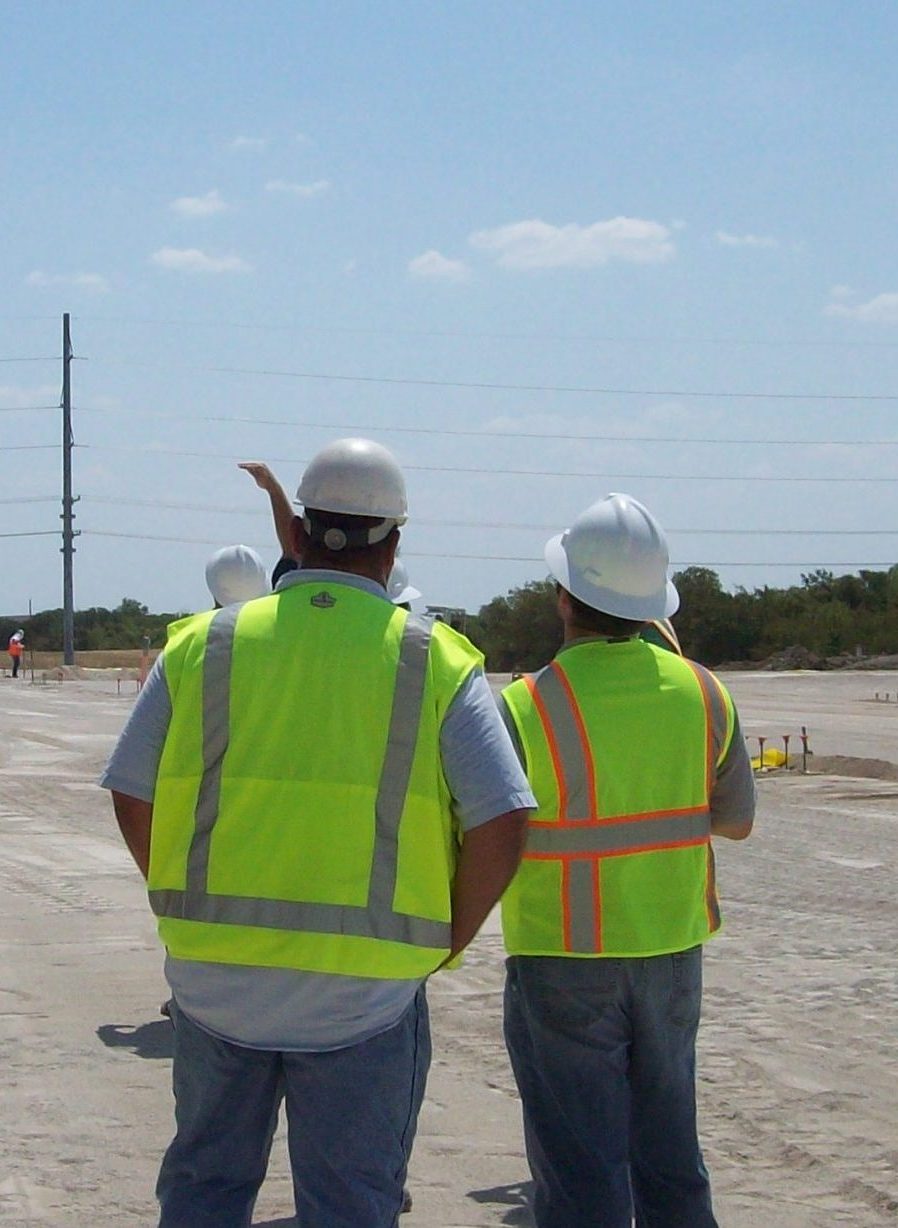 Two construction workers wearing high-visibility vests and hard hats stand on a construction site. One gestures forward while the other looks in the same direction. Power lines and equipment are visible.