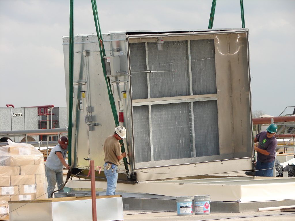 Construction workers installing a large industrial air conditioning unit onto a rooftop using straps and cranes. Various tools and supplies are scattered around the work area.