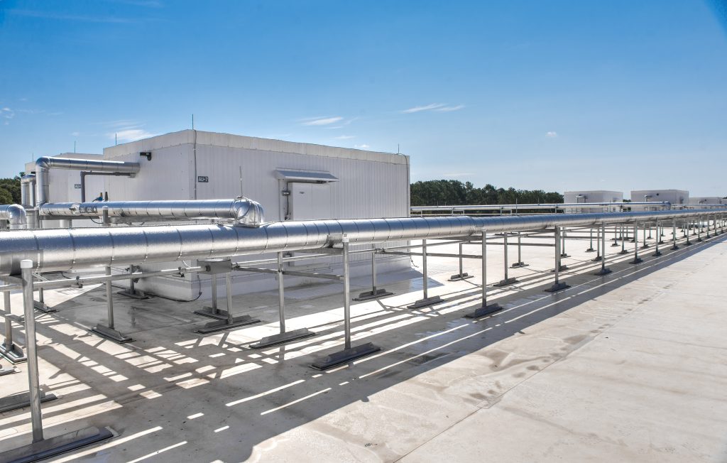 Rooftop view of an industrial facility showing a white building and extensive network of exposed metal pipes under a clear blue sky.