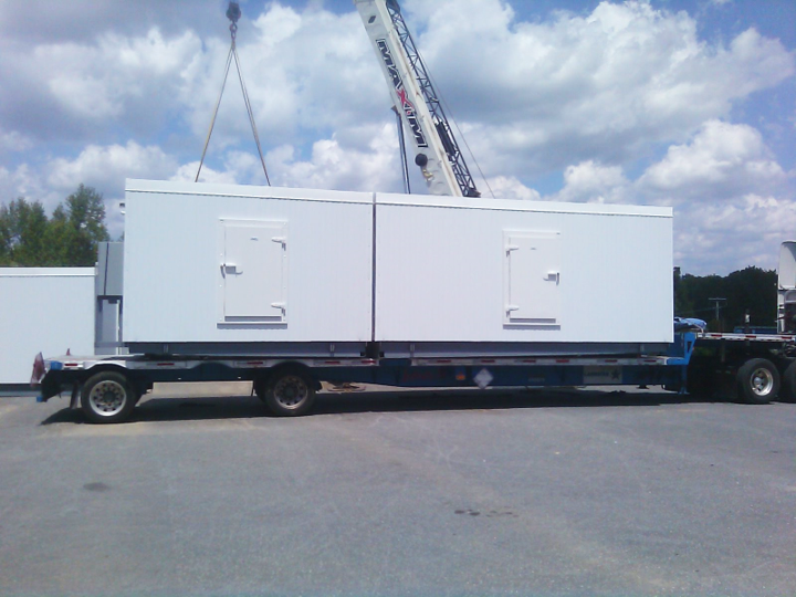 A large white modular building is being transported on a flatbed trailer, with a crane visible in the background against a partly cloudy sky.