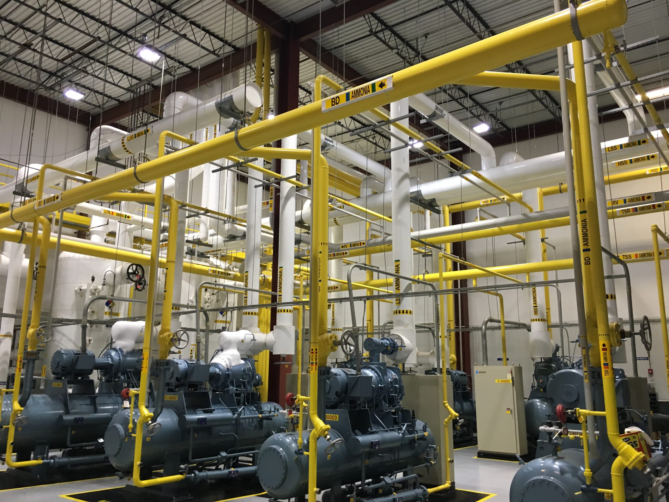 Industrial facility interior with large yellow metal pipes, storage tanks, blue machinery, and electrical control panels. The ceiling features structural beams and lighting fixtures.