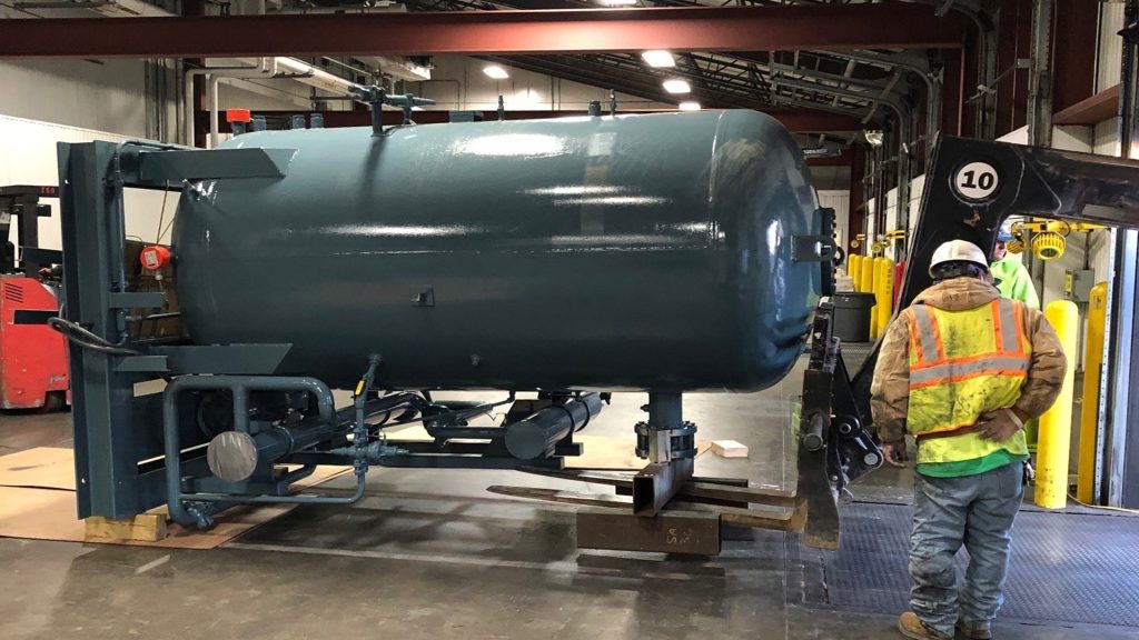 A worker wearing a hard hat and safety gear stands next to a large horizontal cylindrical tank being transported inside a facility, with machinery and industrial equipment visible in the background.